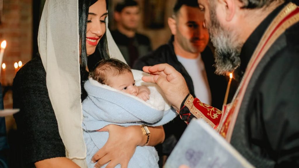Intimate orthodox baptism scene with priest, baby, and family members indoors.