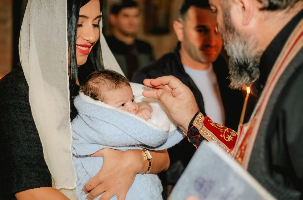 Intimate orthodox baptism scene with priest, baby, and family members indoors.